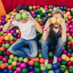 Dwójka dzieci, chłopiec i mężczyzna siedzące w basenie kulkowym i trzymające kulki w rękach zasłaniając nimi oczy.