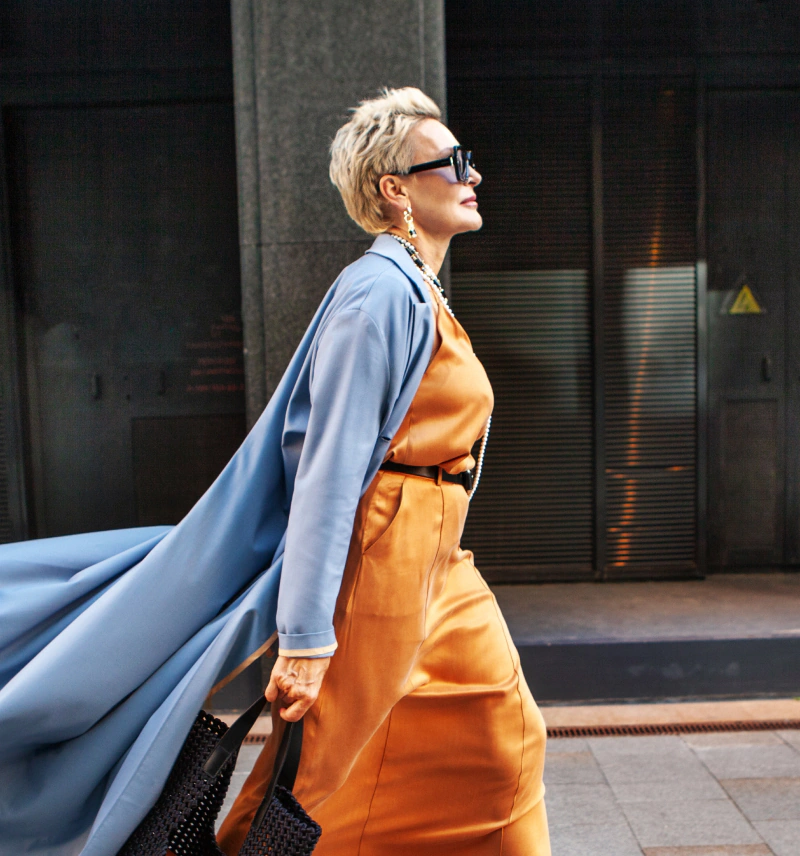 Modnie ubrana kobieta w średnim wieku w błękitnym płaszczu i pomarańczowej sukience idąca chodnikiem przed centrum handlowym.
